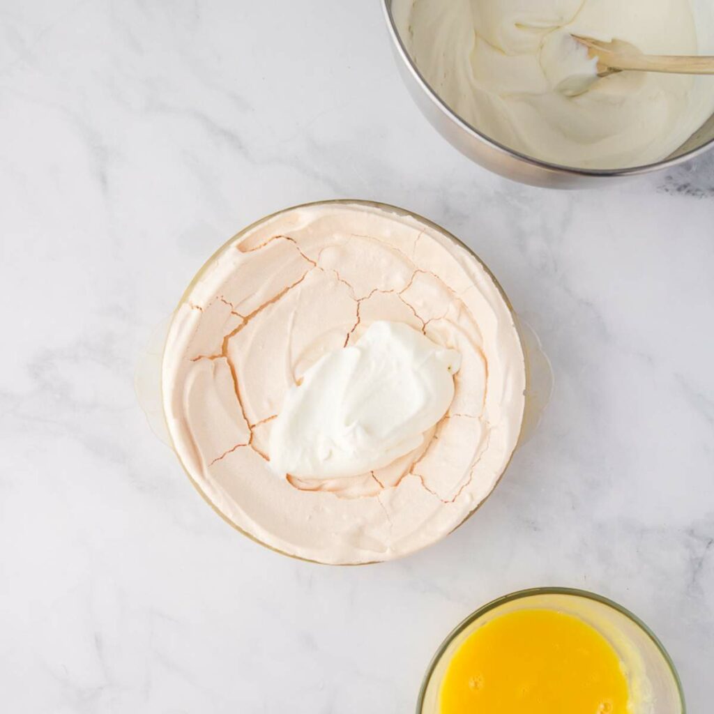 Bowl of pavlova with cracks and a dollop of whipped cream on top, next to a bowl of lemon curd and a metal bowl with cream and a spatula on a marble surface.