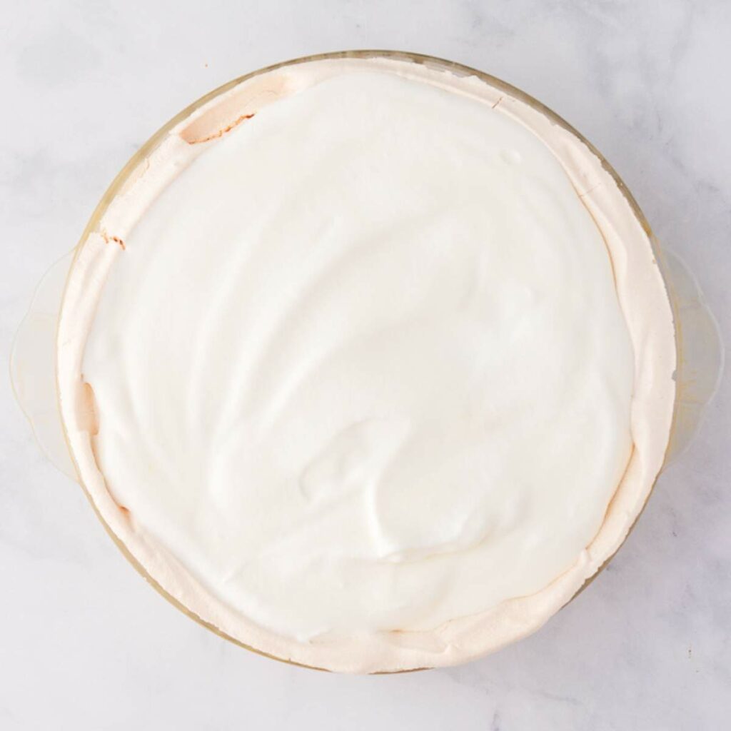 A pie in a glass dish topped with a smooth layer of white whipped cream, viewed from above on a light surface.