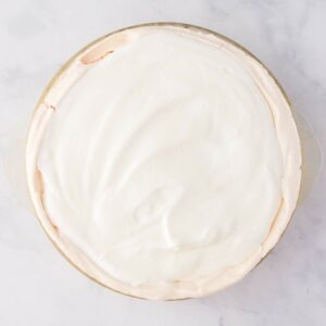 A pie in a glass dish topped with a smooth layer of white whipped cream, viewed from above on a light surface.