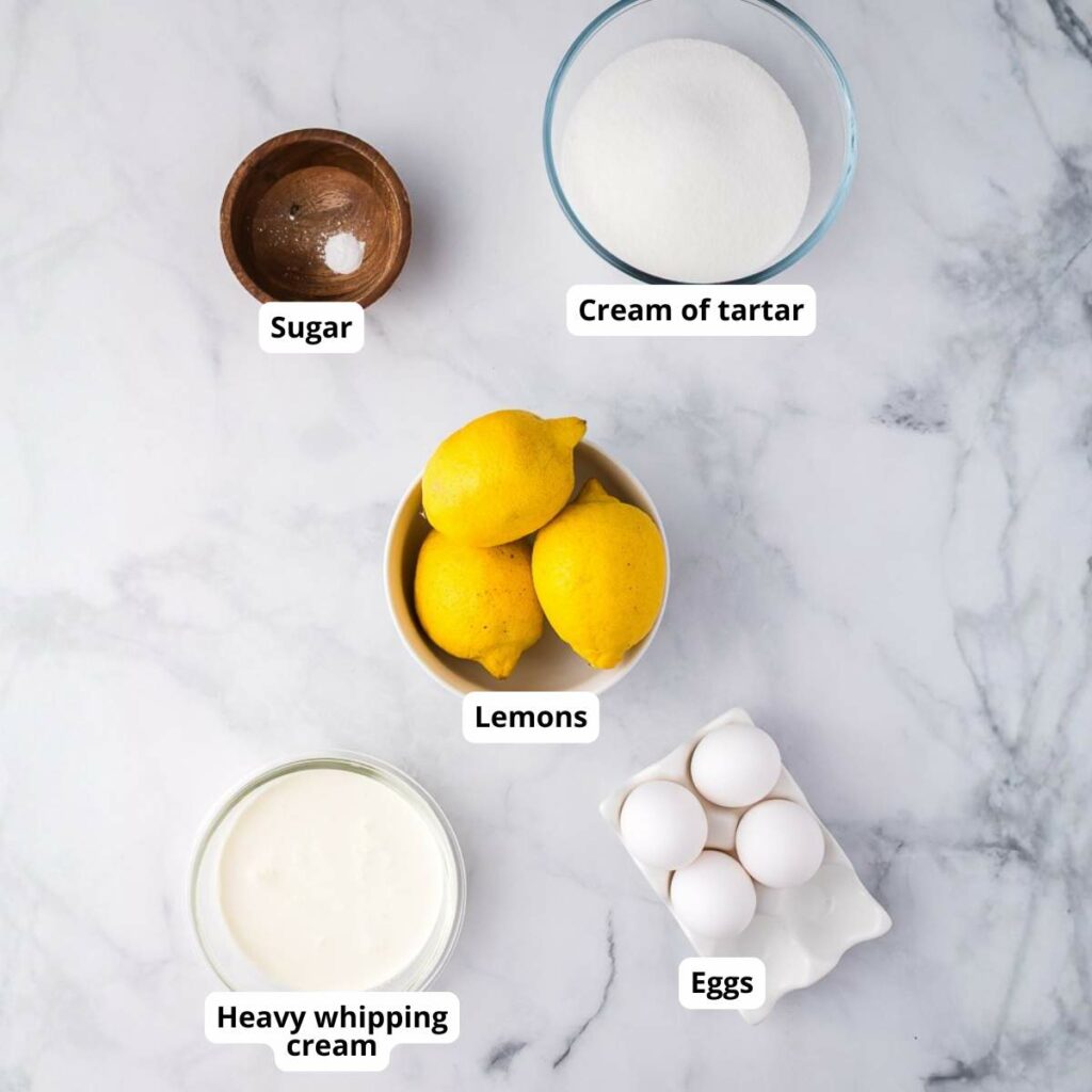 Five labeled bowls on a marble surface containing sugar, cream of tartar, lemons, heavy whipping cream, and eggs.