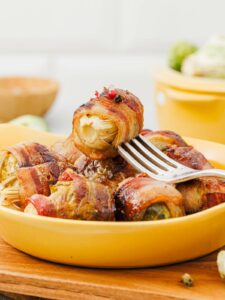 A fork holds a piece of bacon-wrapped brussels sprout above a yellow dish filled with several more bacon-wrapped brussels sprouts.