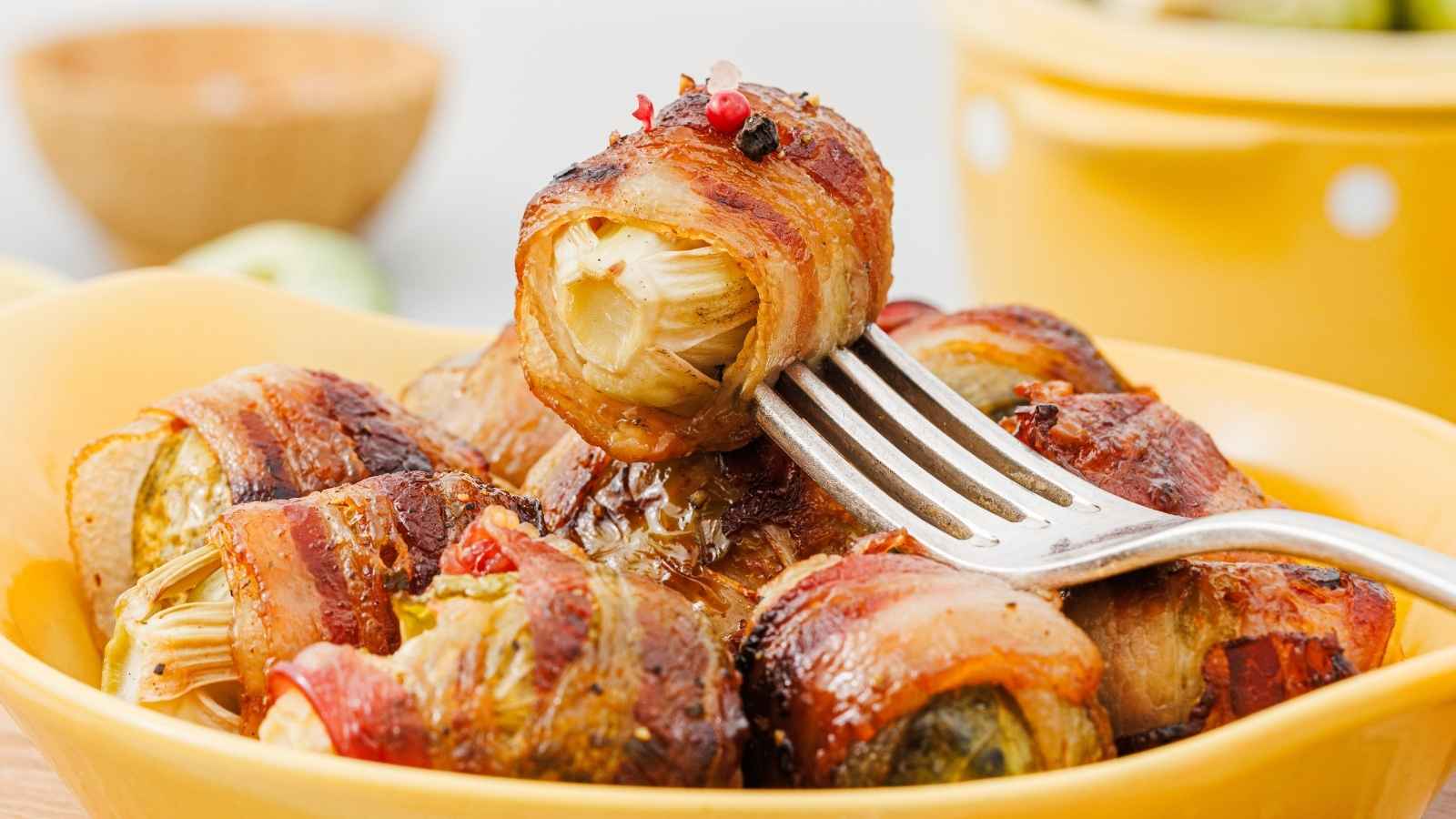 A fork holds a bacon-wrapped brussels sprout above a yellow dish filled with more bacon-wrapped brussels sprouts.