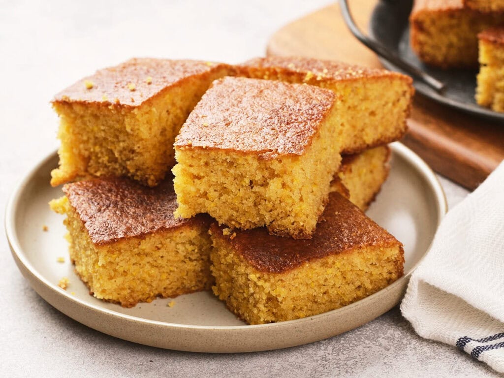 A plate with six square pieces of golden-brown cornbread, stacked neatly on a light-colored surface.