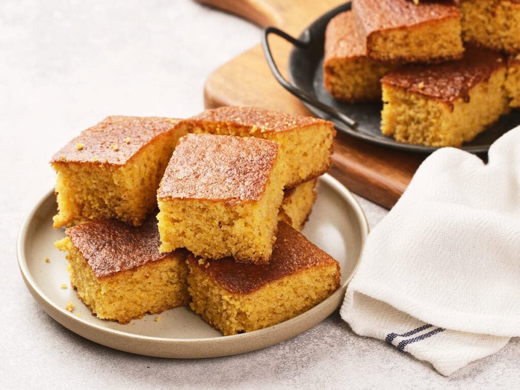 A plate of six square pieces of golden-brown cornbread, with more pieces on a tray in the background and a white napkin beside the plate.