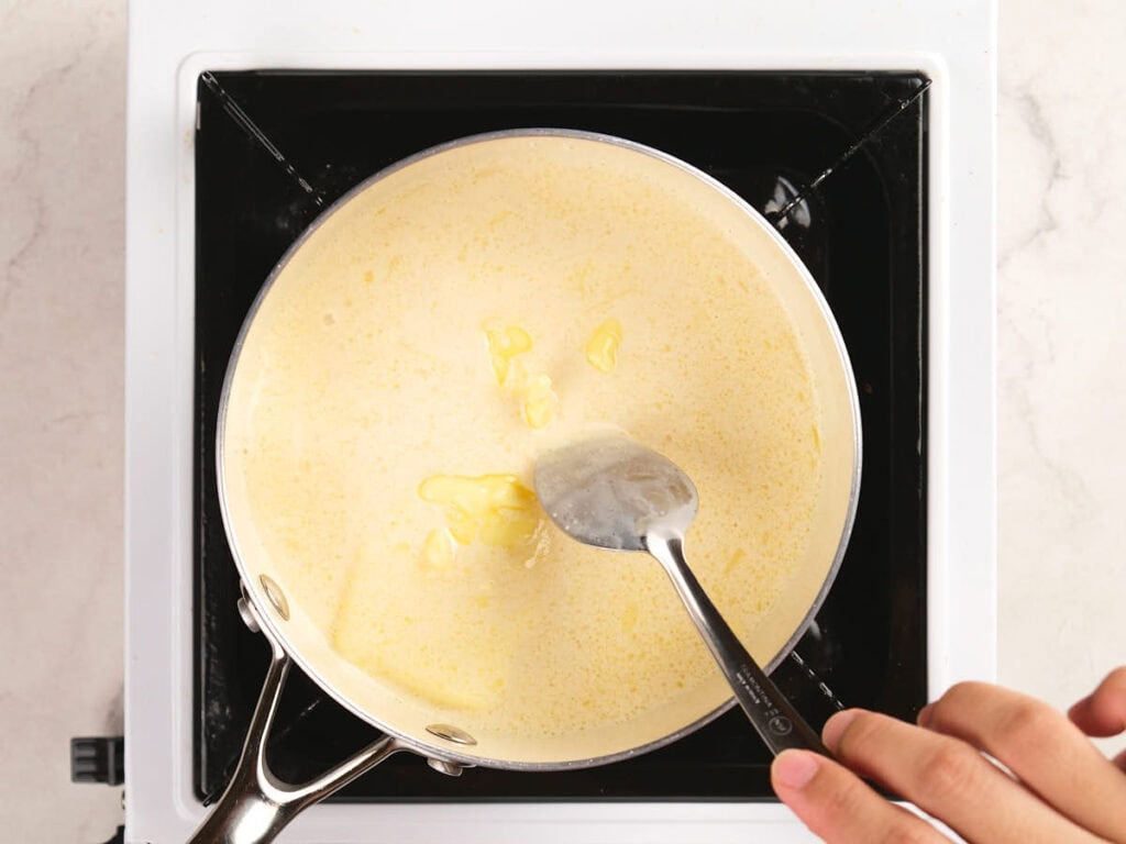 A hand uses a spoon to stir a creamy yellow mixture in a saucepan on a stovetop.