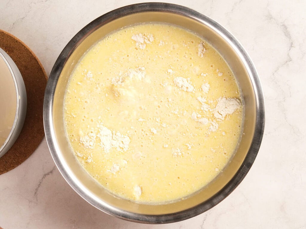 A metal mixing bowl containing a yellow batter with visible lumps and patches of unmixed white flour on the surface.