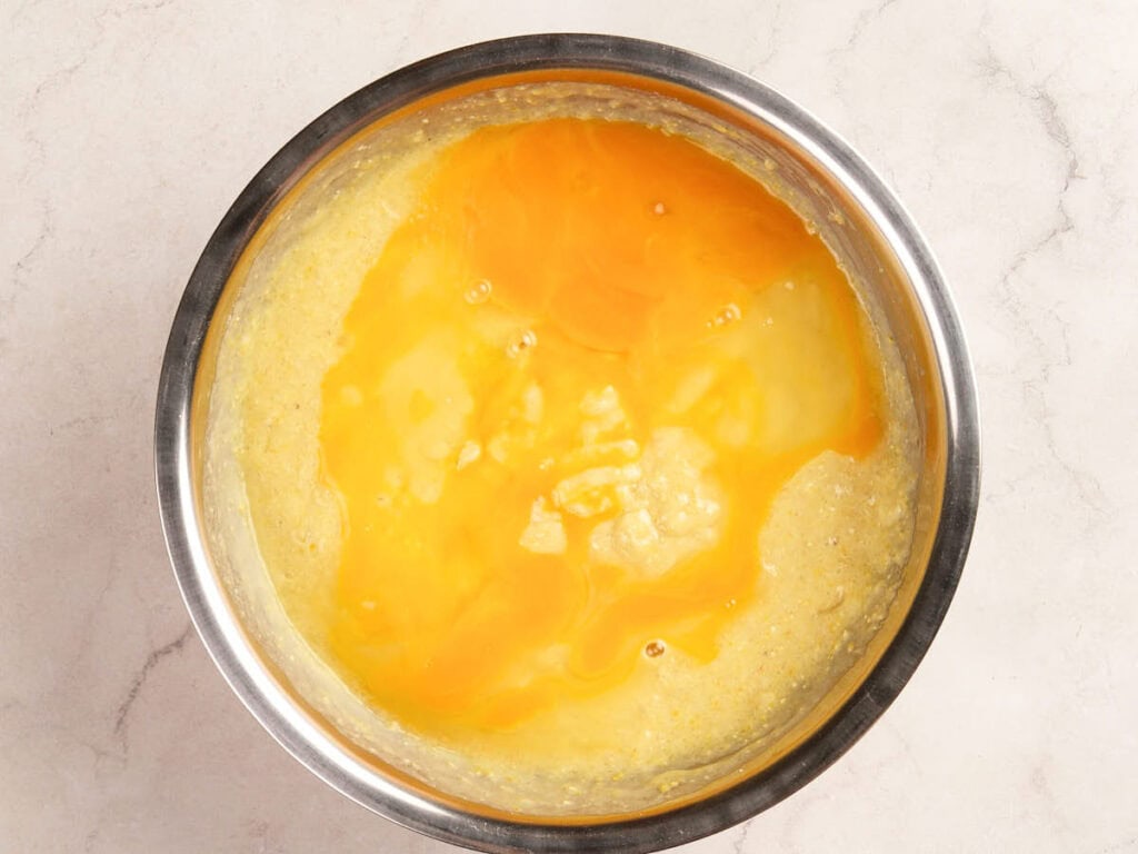 A metal mixing bowl contains a yellowish batter with raw eggs cracked on top, ready to be mixed.