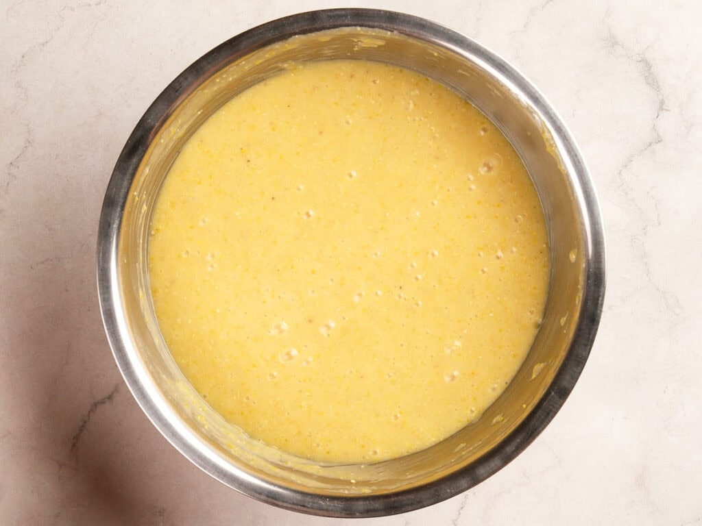 A metal mixing bowl containing a yellow cornbread batter sits on a light marble countertop.