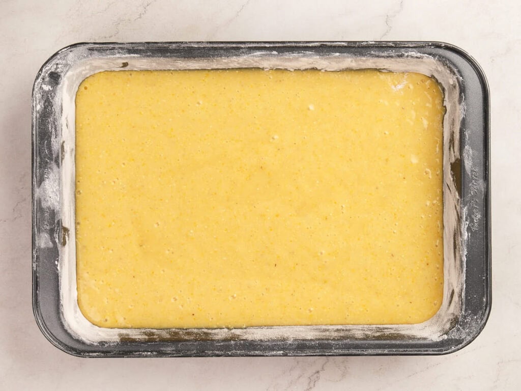 Rectangular metal baking pan filled with uncooked yellow cornbread batter, lightly dusted with flour around the edges, on a light marble surface.