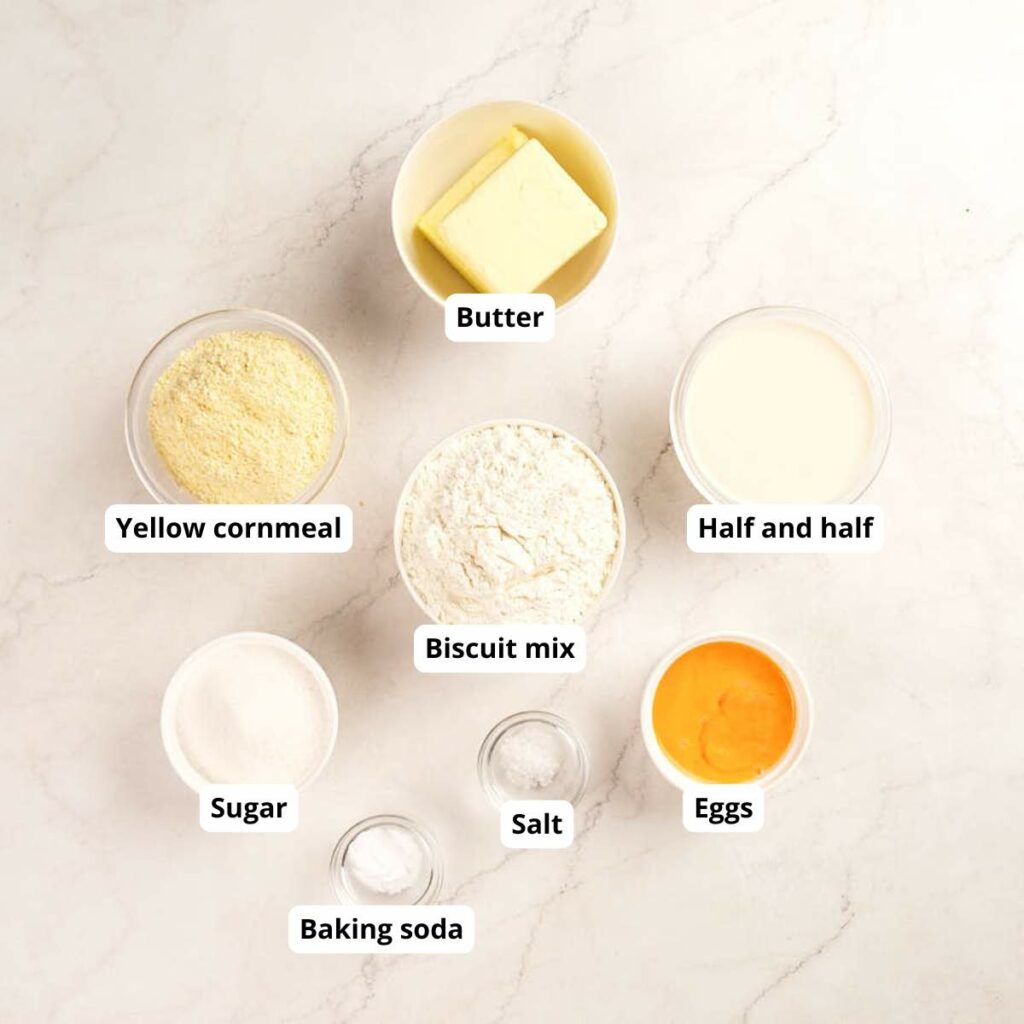 Seven labeled bowls and containers on a light surface, each holding an ingredient: butter, yellow cornmeal, half and half, biscuit mix, sugar, salt, baking soda, and eggs.