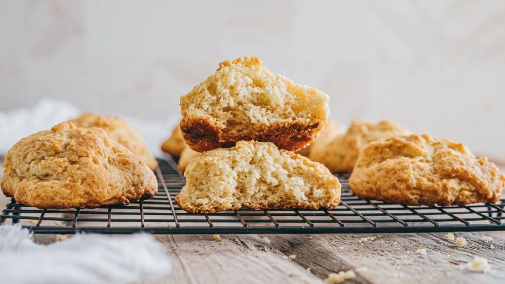 Golden brown drop biscuits cooling on a wire rack, with one biscuit split open to show its soft, fluffy interior.