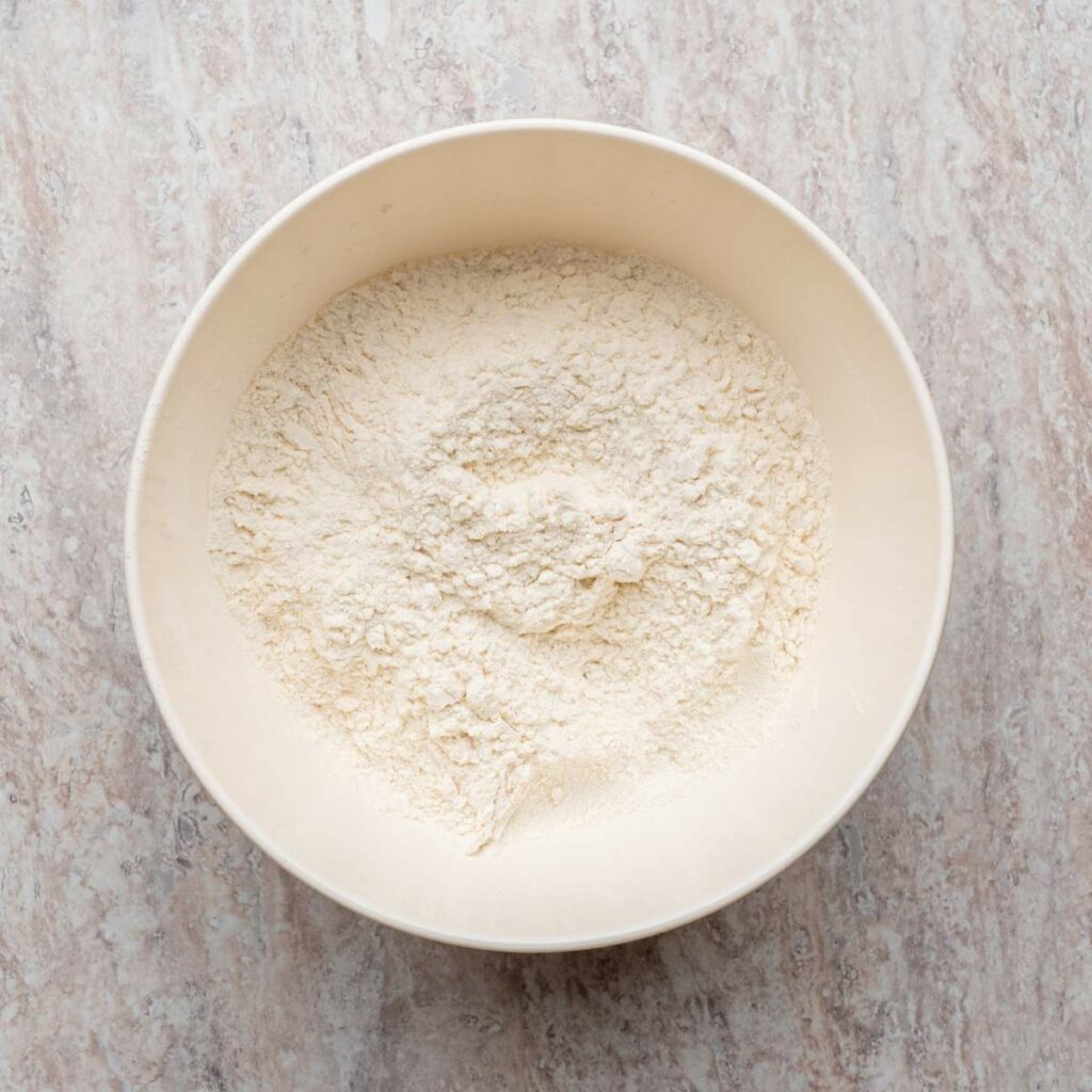 A white mixing bowl filled with flour sits on a light-colored marble countertop.