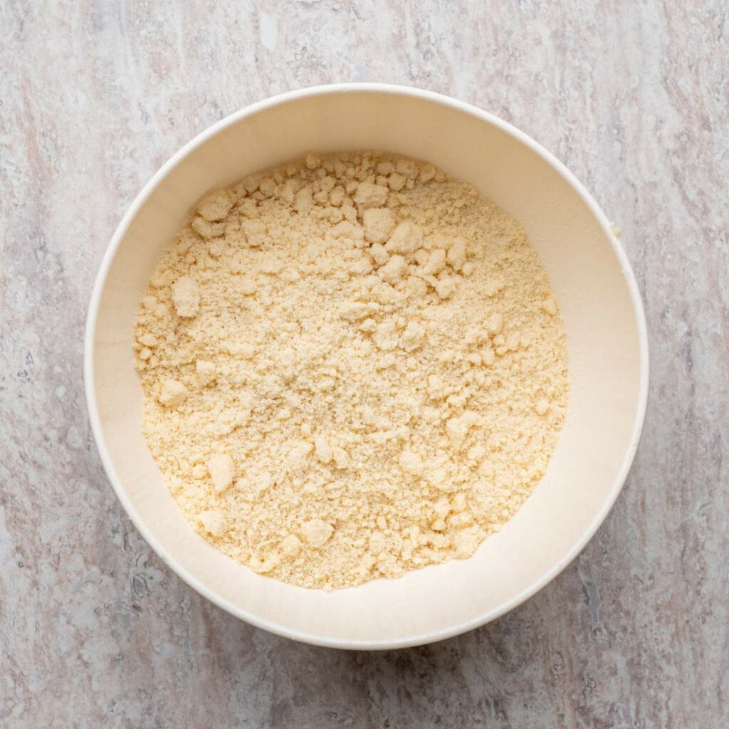 A white bowl filled with a crumbly mixture of flour and butter sits on a light-colored, textured countertop.