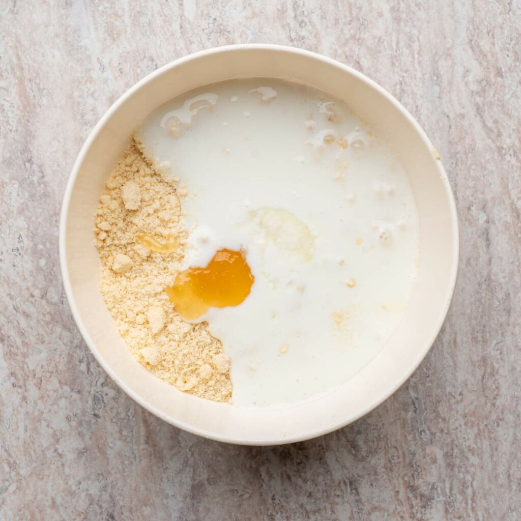 A bowl containing flour, milk, and a cracked egg on a light-colored countertop, with the ingredients unmixed.