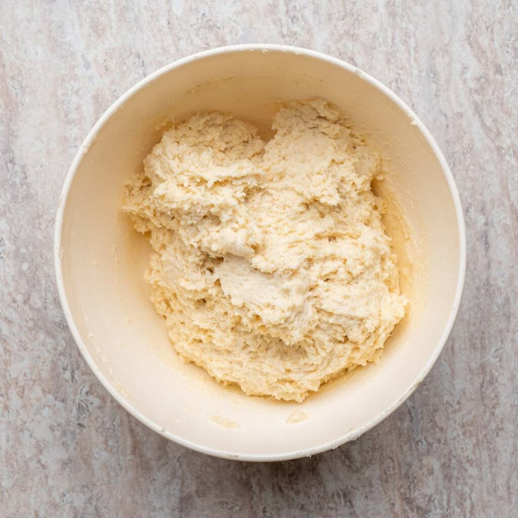 A white mixing bowl filled with raw dough sits on a light-colored, textured countertop.
