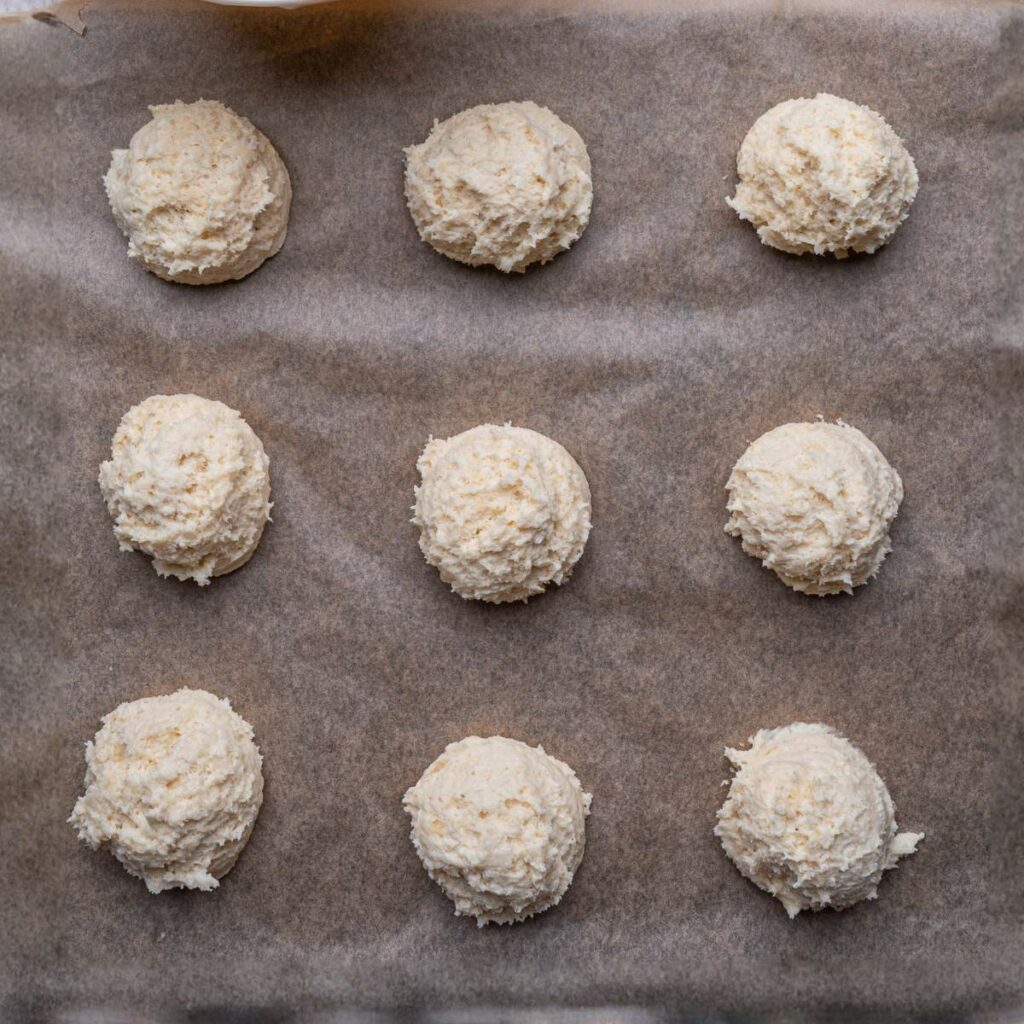Nine evenly spaced scoops of unbaked biscuit dough on a parchment-lined baking sheet.