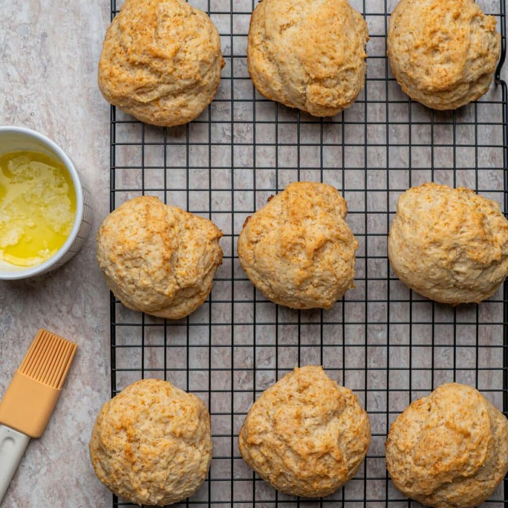Nine golden brown biscuits cool on a wire rack next to a bowl of melted butter and a pastry brush.