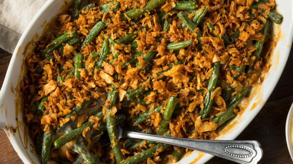 Green bean casserole topped with crispy fried onions in a white baking dish, with a serving spoon inside.
