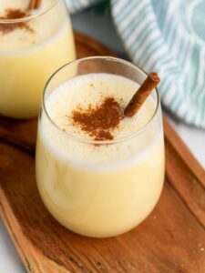 A glass of creamy eggnog topped with ground cinnamon and a cinnamon stick, placed on a wooden tray.