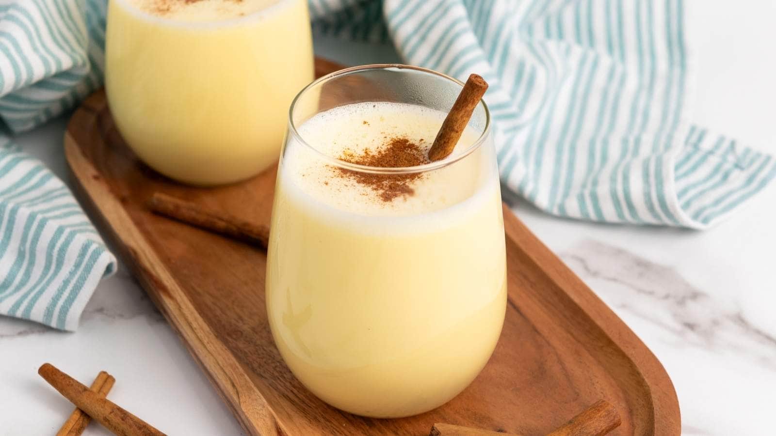 Two glasses of eggnog are on a wooden tray, garnished with cinnamon sticks and sprinkled with ground cinnamon.