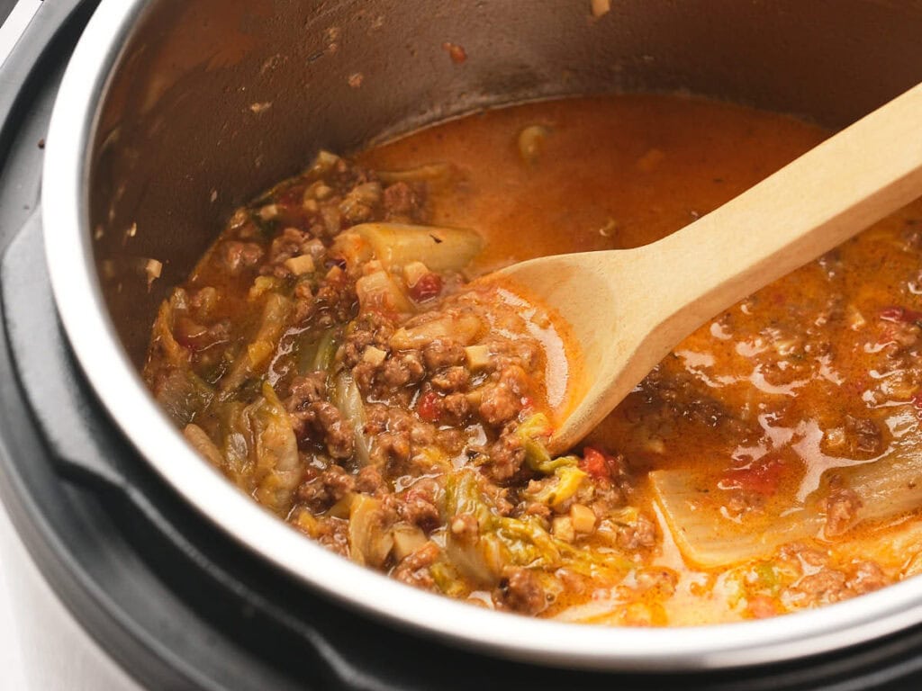 A wooden spoon stirs a pot of cooked ground meat, vegetables, and sauce inside an electric pressure cooker.