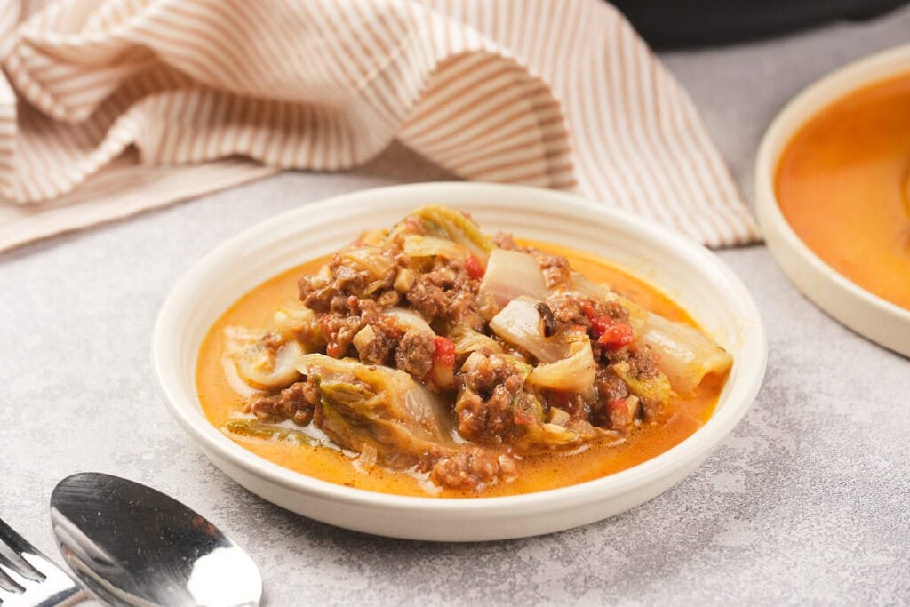 A white plate with a stew containing ground meat, cabbage, onions, and red peppers in a reddish broth sits on a light surface near utensils.