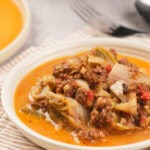 A shallow white bowl filled with cabbage roll soup containing ground meat, chopped cabbage, tomatoes, and broth, placed on a striped cloth.