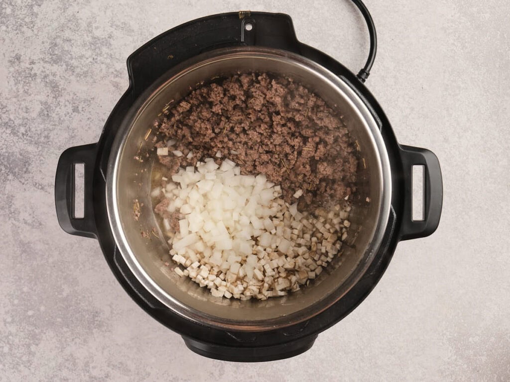 Ground beef, diced onions, and chopped celery cooking in an electric pressure cooker on a light-colored countertop.