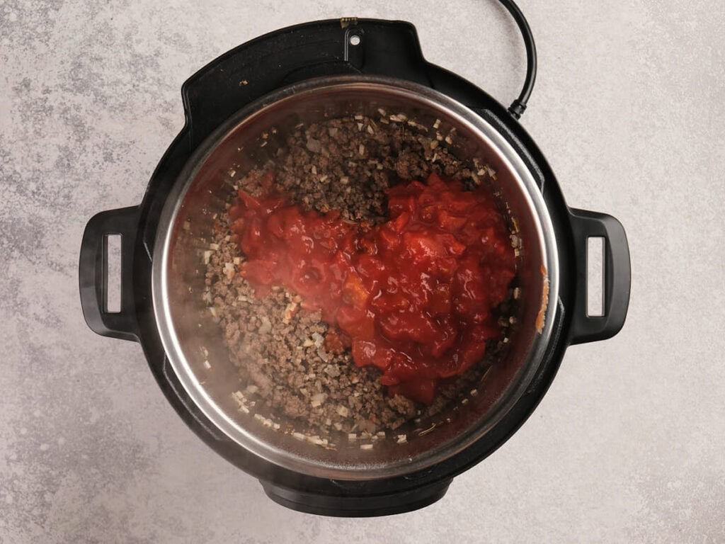 Ground meat, diced onions, and chopped tomatoes cooking in an electric pressure cooker on a light gray countertop.