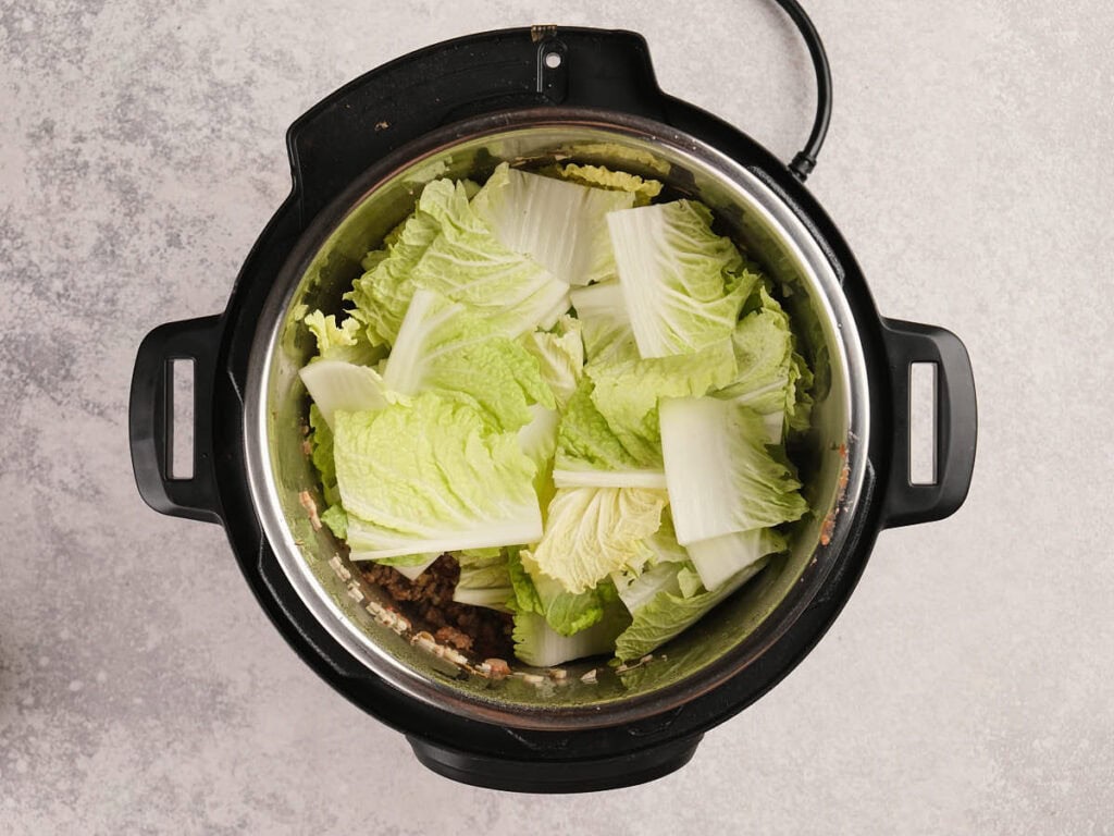 Chopped napa cabbage in an electric pressure cooker with visible ground meat underneath on a light countertop.