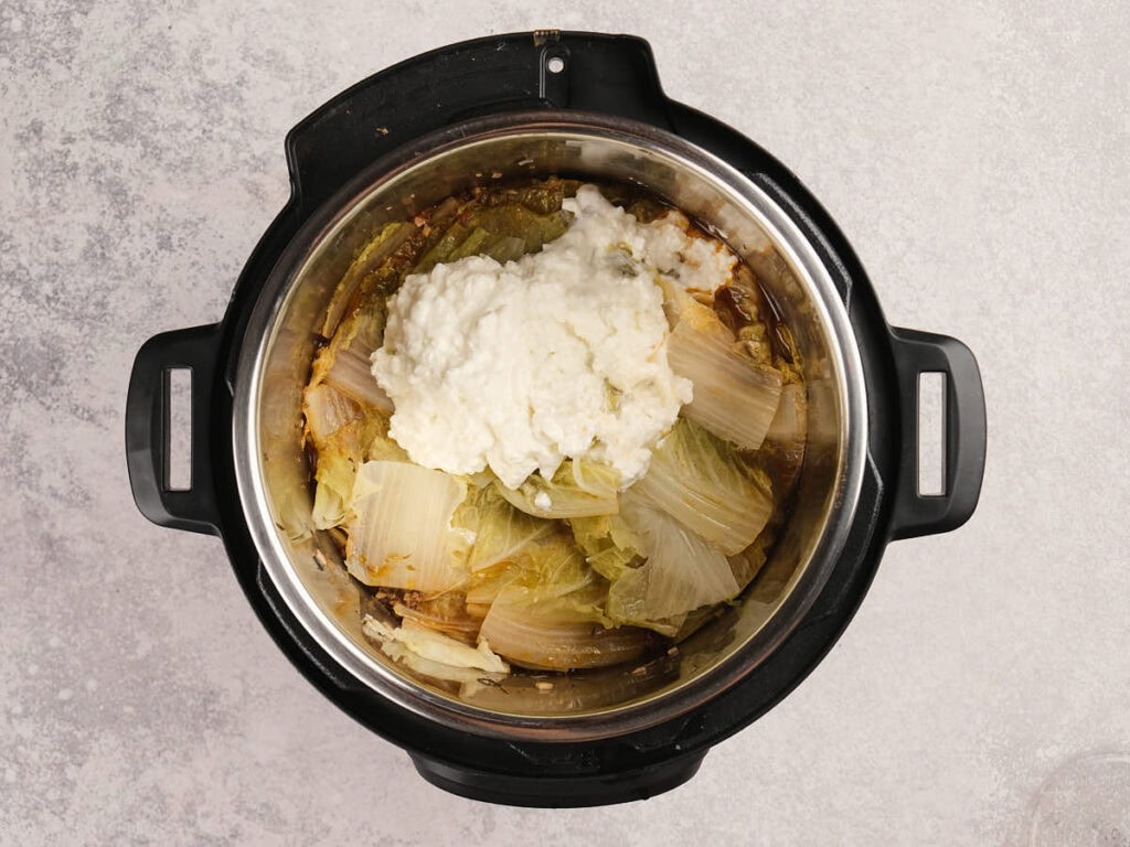 An overhead view of cooked cabbage leaves and a dollop of cottage cheese in a stainless steel pot on a light countertop.