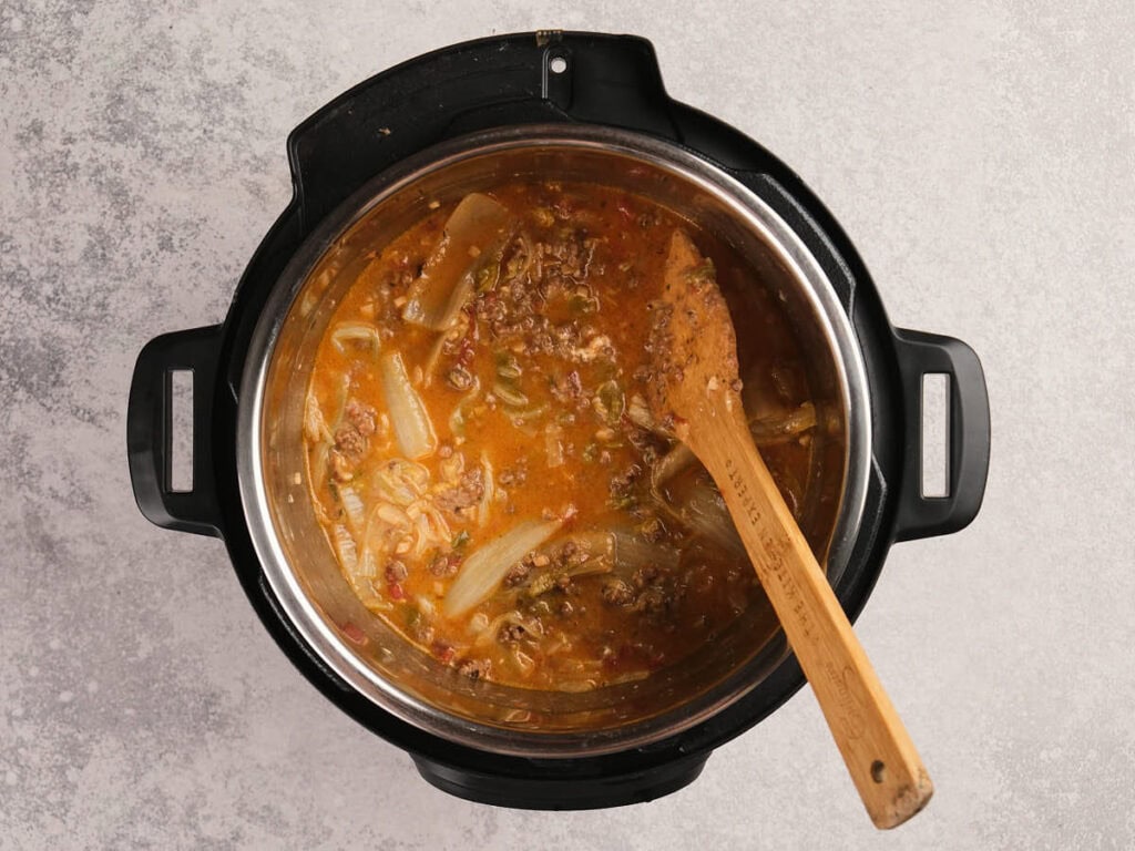 A top-down view of a stew with ground meat and vegetables in an Instant Pot, stirred with a wooden spoon.