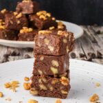 Three pieces of chocolate fudge with walnuts are stacked on a white plate, with more fudge pieces and walnuts in the background.