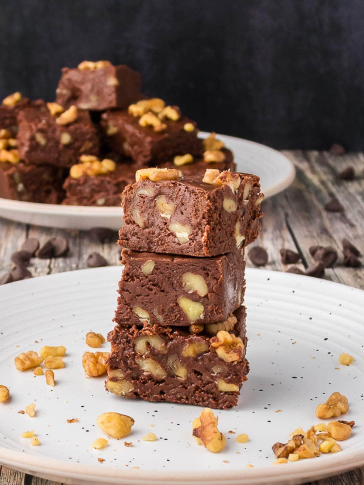 Three pieces of chocolate fudge with walnuts are stacked on a white plate, with more fudge pieces and walnuts in the background.