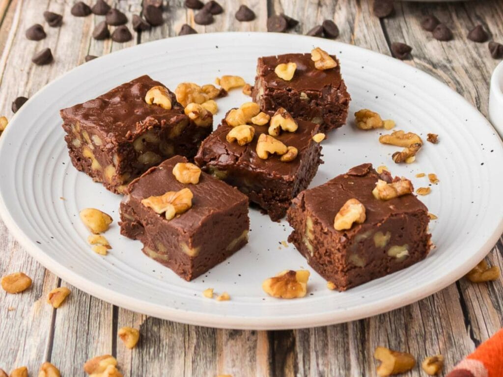 Five squares of chocolate fudge topped with walnut pieces are arranged on a white plate, with extra walnuts and chocolate chips scattered around.