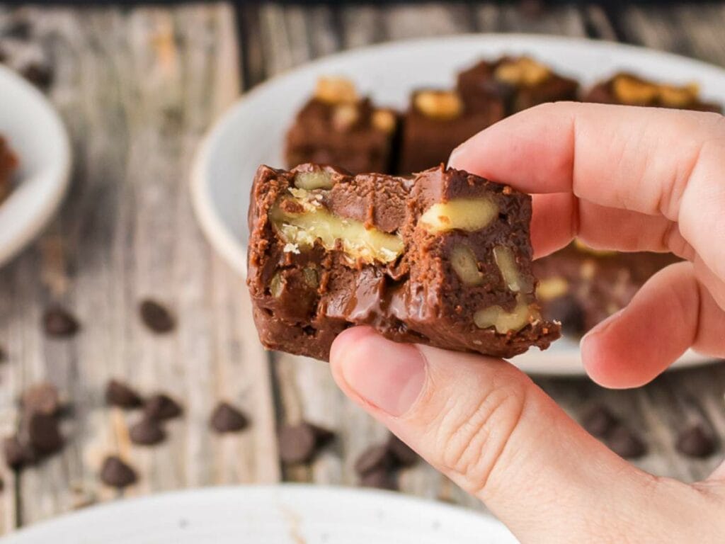 A hand holding two pieces of chocolate fudge with visible walnuts, with more fudge pieces on a plate in the background.