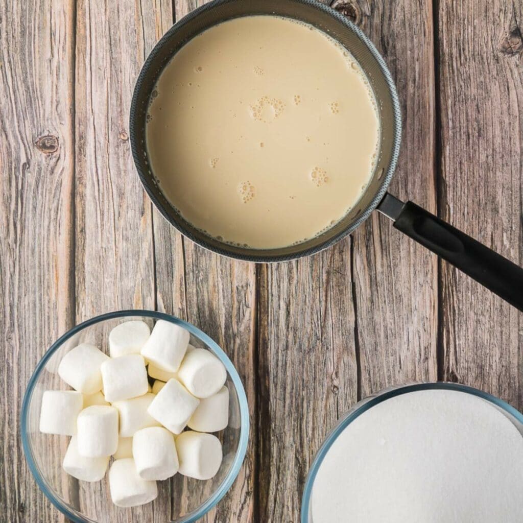 A saucepan with a creamy liquid next to bowls of marshmallows and sugar on a wooden surface.