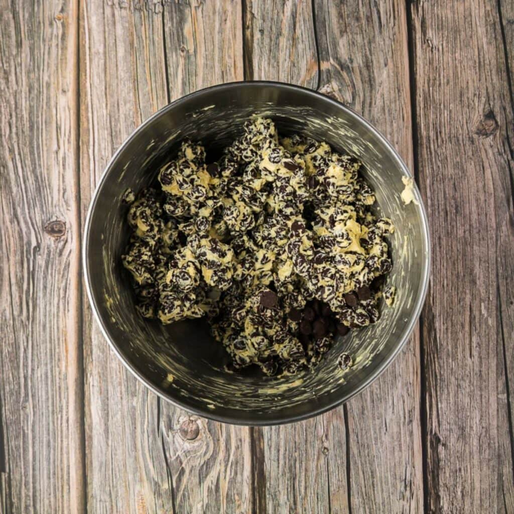 A metal bowl filled with chocolate chip fudge mixture sits on a wooden surface.
