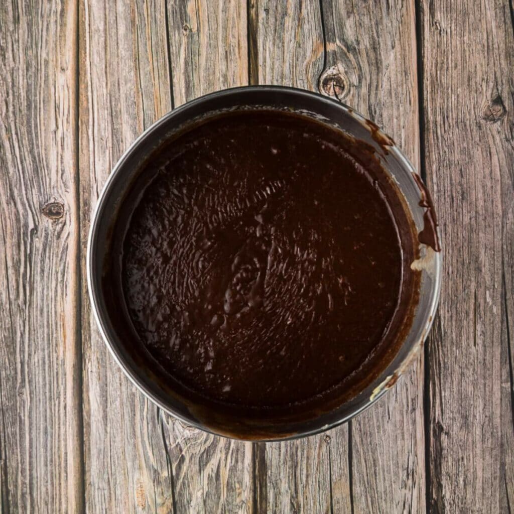 A round metal bowl filled with chocolate fudge batter sits on a rustic wooden surface.