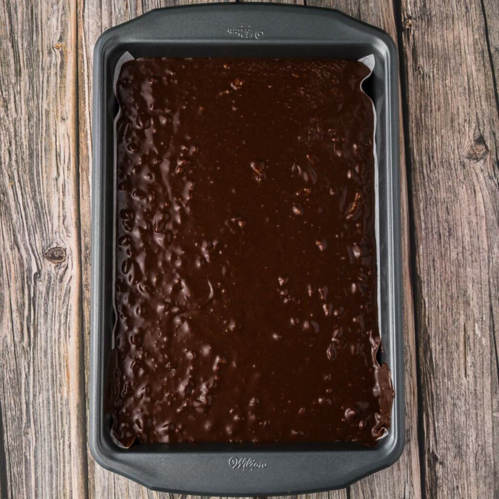 A rectangular metal pan filled with chocolate fudge batter sits on a wooden surface.