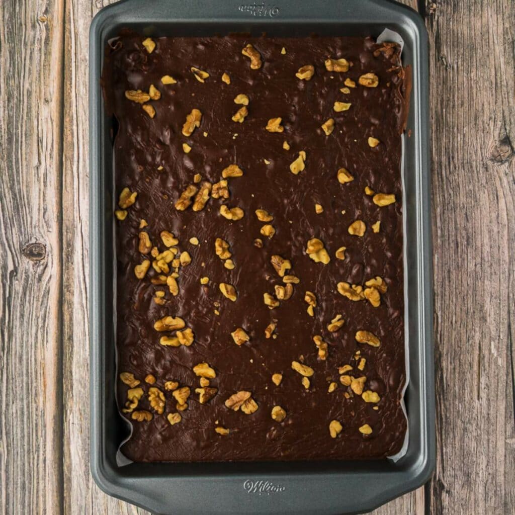 A rectangular baking pan filled with chocolate fudge topped with scattered walnut pieces, placed on a wooden surface.