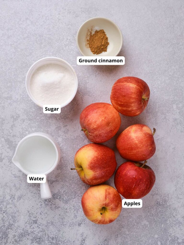 Top-down view of apples, a cup of water, a bowl of sugar, and a small dish of ground cinnamon on a light surface, each labeled with text.