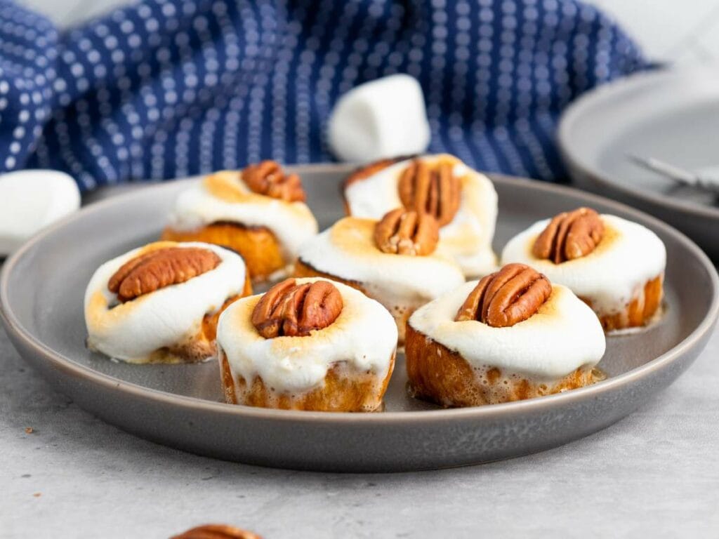 A gray plate holds seven sweet potatoes topped with toasted marshmallows and pecan halves; a blue patterned cloth is in the background.