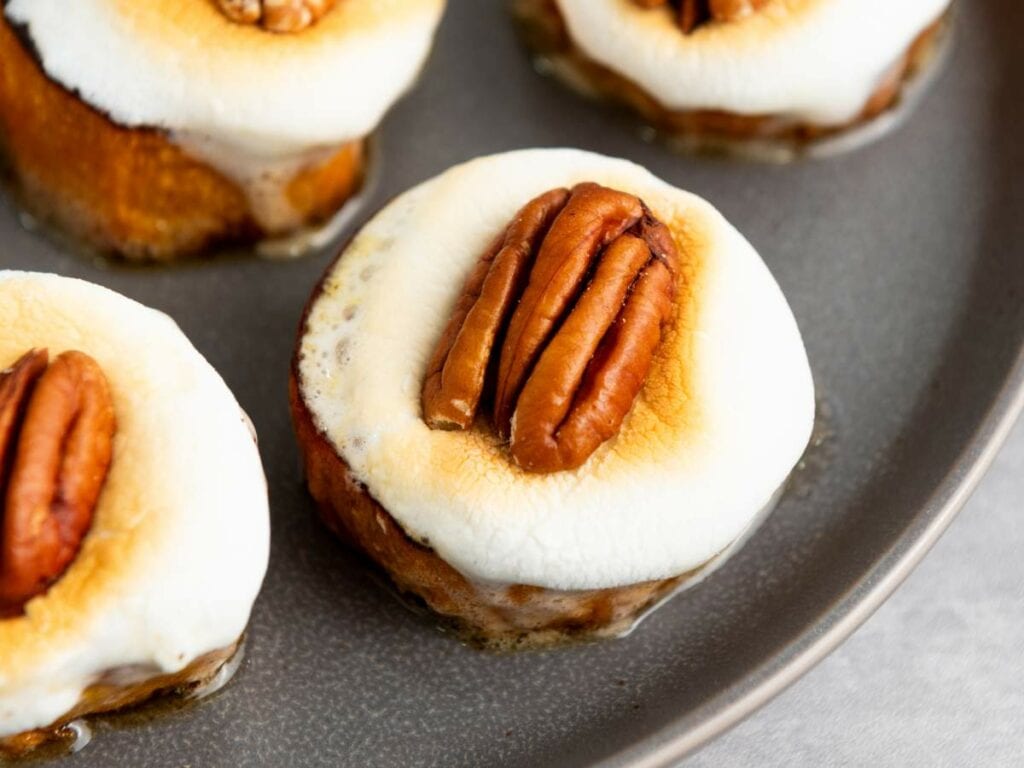 Close-up of sweet potato rounds topped with melted marshmallows and pecan halves, arranged on a gray plate.
