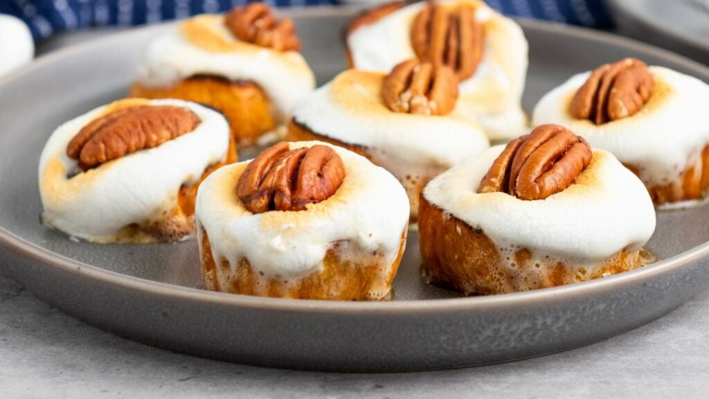 Six sweet potato pieces topped with melted marshmallows and pecan halves are arranged on a round gray plate.