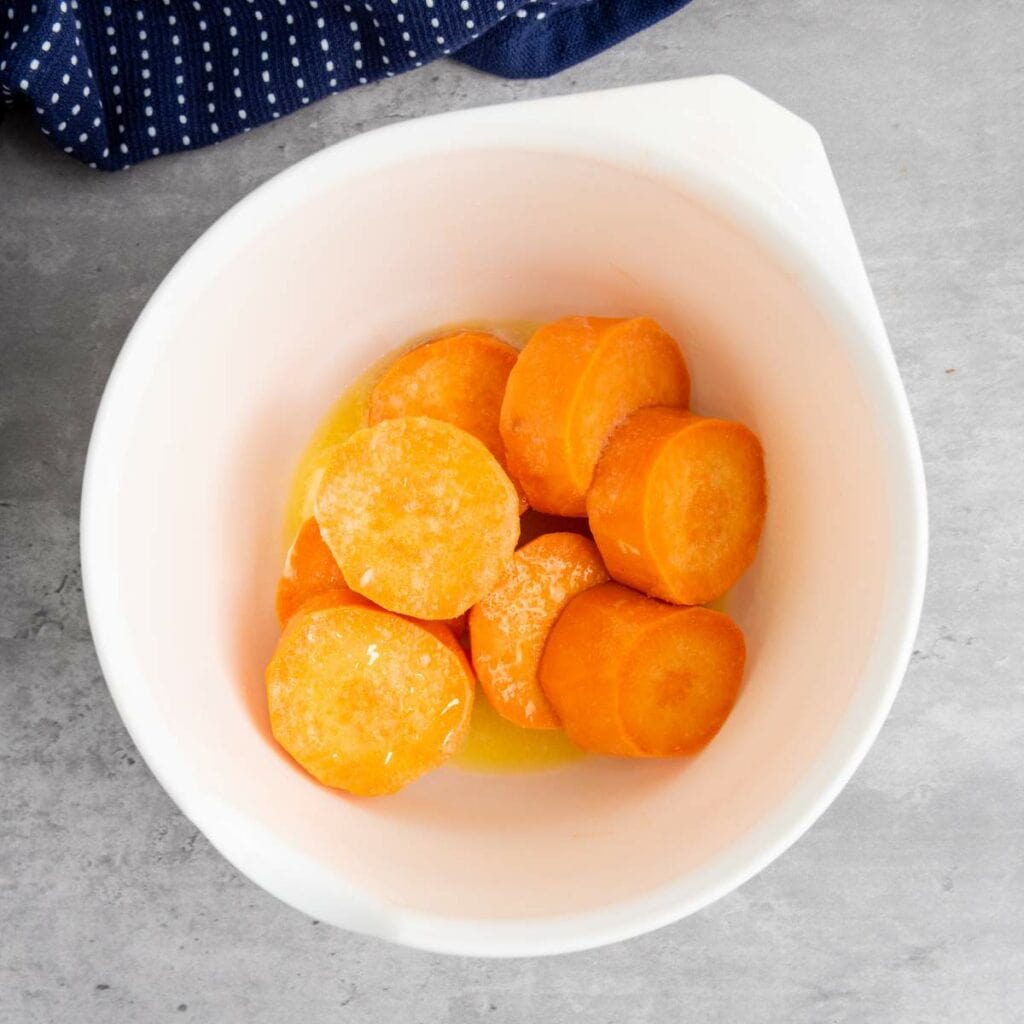 Sliced sweet potatoes coated in melted butter in a white mixing bowl on a gray countertop.