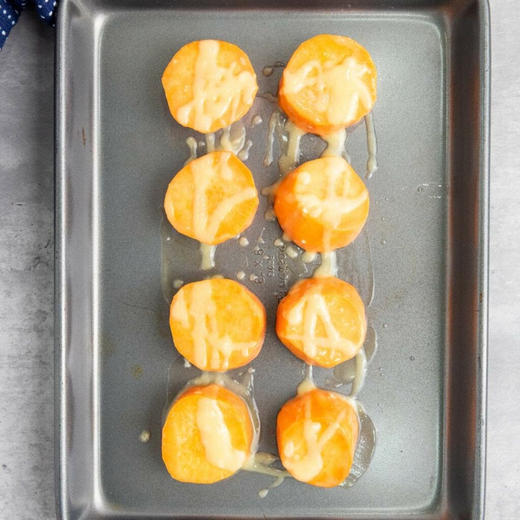 Eight round sweet potato slices with a drizzle of sauce are arranged in two vertical rows on a metal baking sheet.