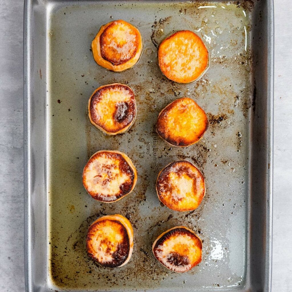 Eight roasted sweet potato rounds on a metal baking sheet with browned edges and visible seasoning.