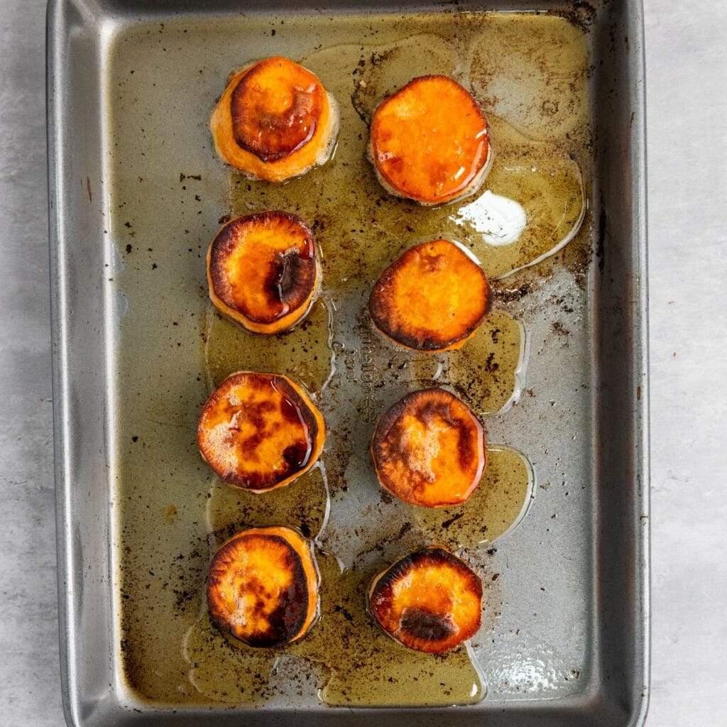 Eight roasted sweet potato rounds on a metal baking sheet, with browned edges and some oil pooled around them.