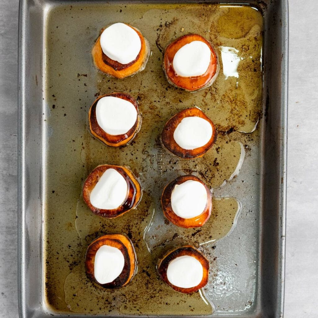 A baking tray with eight roasted sweet potato rounds, each topped with a marshmallow, arranged in two rows.